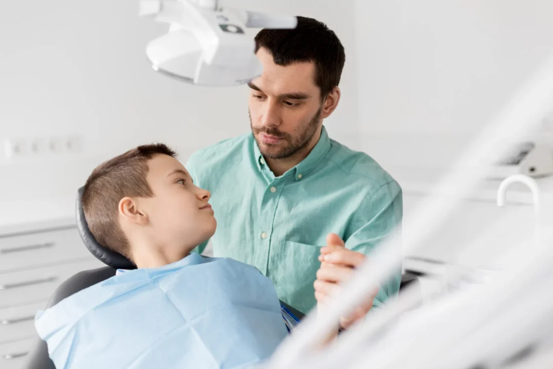 A dental professional in a light green shirt comforts a child seated in a dental chair, covered with a blue bib in a clinic setting.