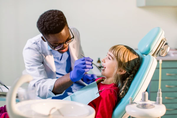 African-American ethnic black male dentist in blue latex gloves checking condition of teeth of little smiling girl. baby girl in blue dental chair in modern dentistry clinic.