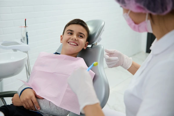 Handsome boy sitting in dentist chair smiles with beautiful toothy smile looking at a pediatric dentist teaching him about oral hygiene during dental appointment in white clean modern medical cabinet