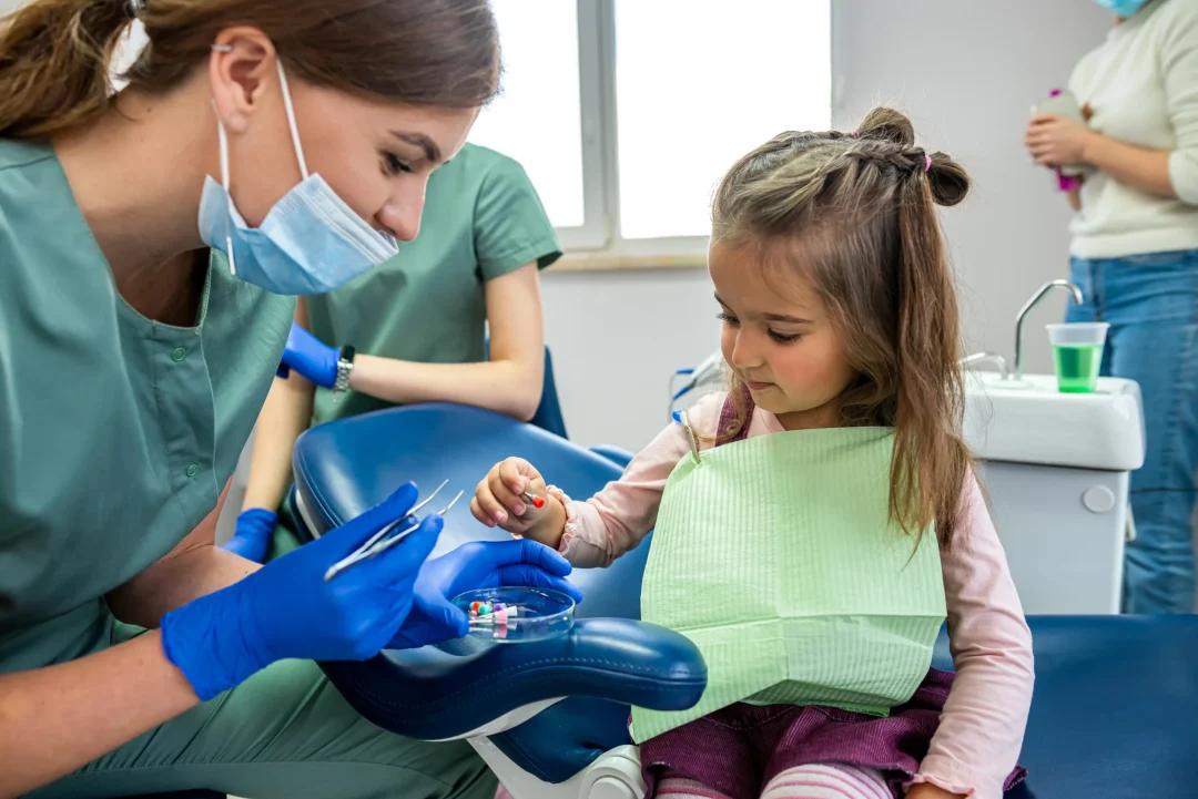 A dental professional in scrubs prepares for a procedure with a young girl, who is seated and wearing a dental bib.