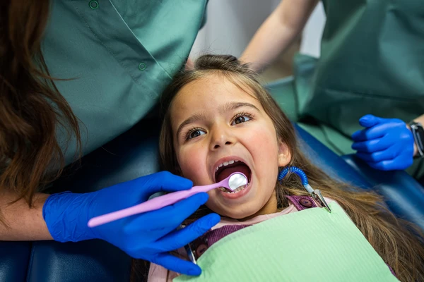 little girl sitting in a dental chair and two female dentists treating baby teeth. Two female dentists entertain a little patient